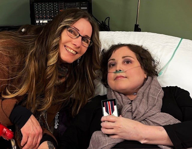 Two women are smiling and facing the camera. One has long brown hair, the other short curly brown hair and is elevated in a hospital type of bed.