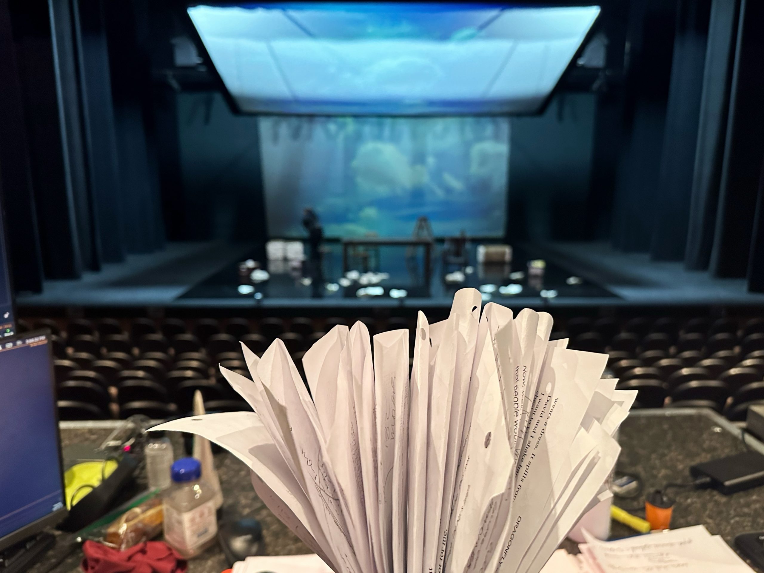 Pages of notes sitting up on desk in front of lit theatre stage